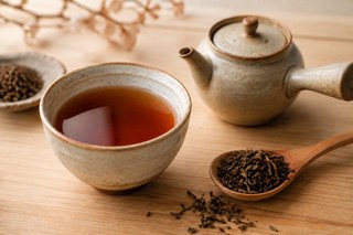 Amber-coloured hojicha in a ceramic cup with a teapot and dark roasted tea leaves on a wooden table.