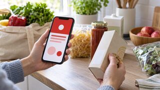 Symbolbild: Eine Person vergleicht auf einer Küchenarbeitsfläche eine abstrakte Warnmeldung auf dem Smartphone mit dem leeren Chargenfeld einer neutralen Verpackung.