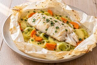 Geöffnetes Backpapierpäckchen mit gegartem weißem Fisch, Kartoffelscheiben, Lauch und Möhren